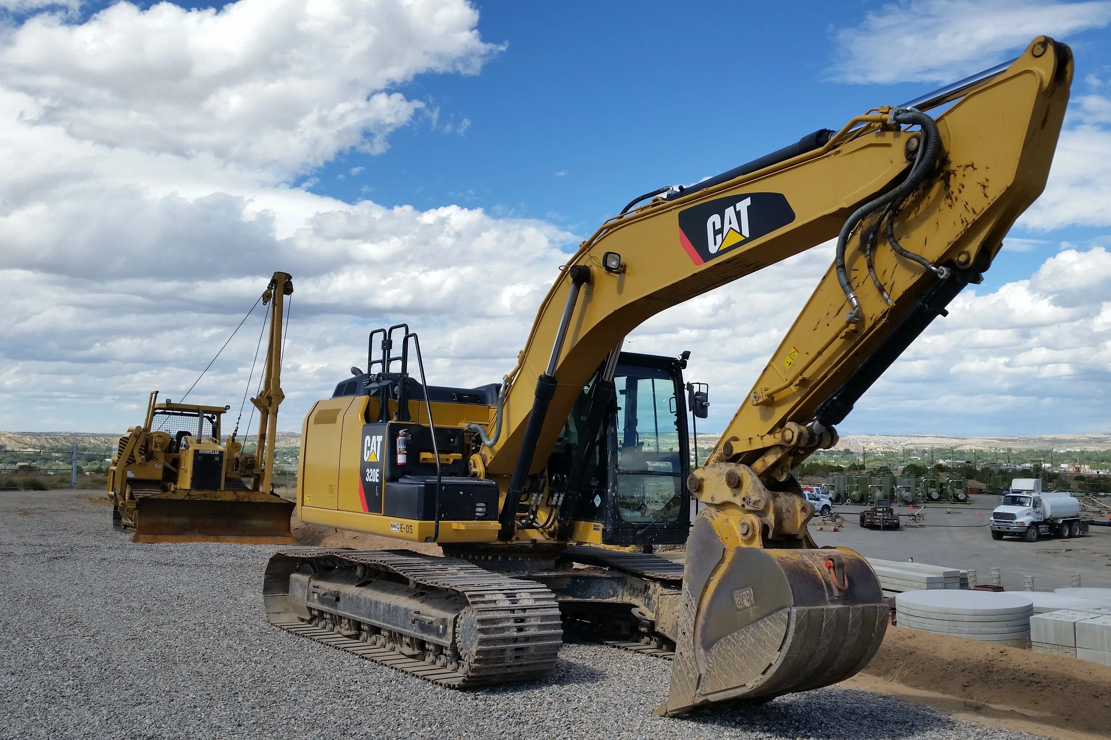 A yellow Cat hydraulic excavator on a gravel pad, a pipelayer behind it, under a wide cloudy sky